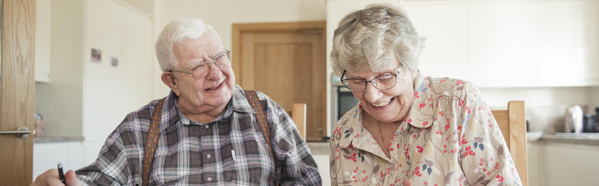 Elderly couple smiling and looking at a newspaper in their kitchen